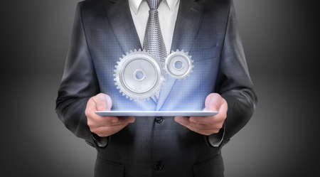 A businessman in a suit holds a glowing tablet with two interlocked metal spur gears hovering above it.の写真素材