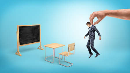 A tiny businessman hanging from a giant hand over blue background with school furniture and a blackboard.の写真素材
