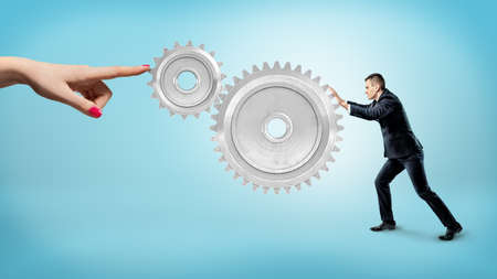 A small businessman pushes at a large gear interlocked with a small one while a female finger stops their movement.の写真素材