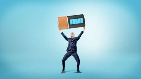 A small happy businessman smiles and holds a huge fully charged battery over his head.の写真素材