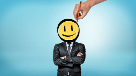 A businessman with crossed hands stands in front view while a giant hand draws a smiley face instead of his head.の写真素材