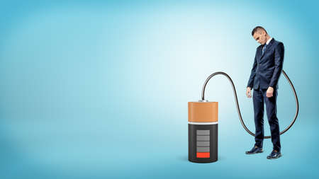 A sad businessman with his head down stands connected by cable to a large empty battery.の写真素材