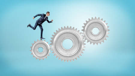 A small businessman runs on large interlocked metal gears on a blue background.の写真素材