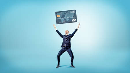 A happy and victorious businessman stands on a blue background and holds up a giant generic credit card.の写真素材