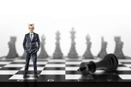 A small businessman with a golden crown on his head stands on a chessboard near a fallen black king.の写真素材