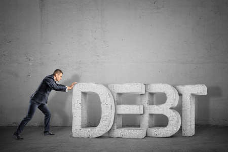 A tiny businessman trying hard to move giant concrete letters forming a word DEBT.の写真素材