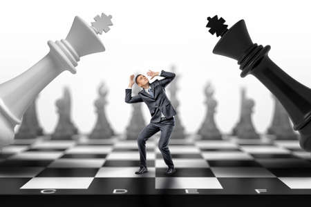 A scared businessman stands between a black and a white chess kings falling on him.の写真素材