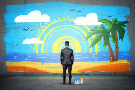 A businessman stands with his back turned and looking at a finished wall picture of exotic isle.の写真素材