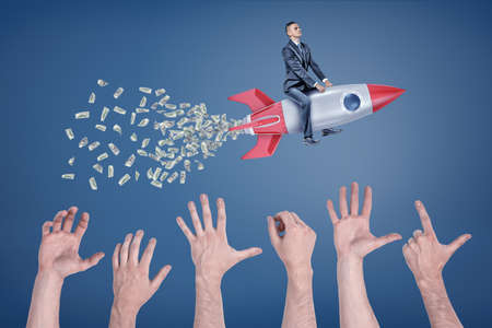 A businessman flies sitting on a rocket that leaves a tail of money with many giant hands trying to catch it.の写真素材