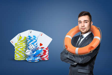 A businessman wears a life buoy on his shoulders and stands near gambling chips, cards and dice.の写真素材