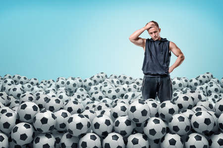 A troubled athletic man stands inside a huge heap of identical football balls.の写真素材