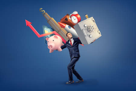 A businessman walks under a heavy weight of many things on his shoulders: a safe box, a piggy bank, skyscraper and an alarm clock.の写真素材
