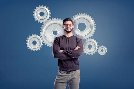 A smart casual man wears glasses and stands in a front view with several large spur gears behind him.の写真素材