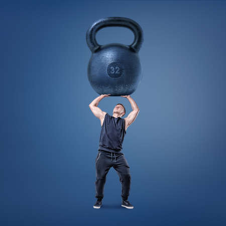 A tiny young muscular man tries to hold a giant iron kettlebell above himself.の写真素材