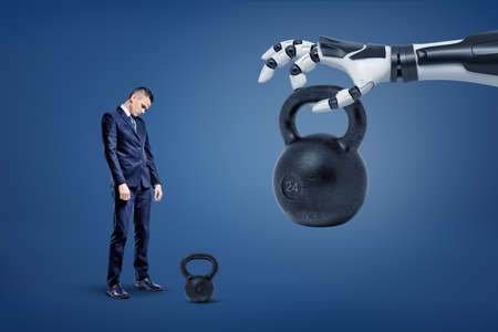 A sad businessman stands near a kettlebell with a robotic hand lifting and giving him another huge one weight.の写真素材