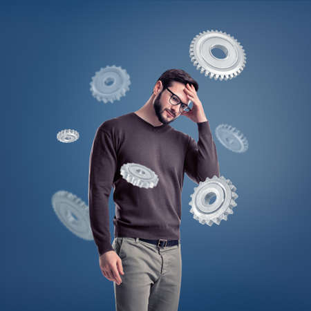A man in glasses stands deep in thought while being surrounded by several large metal spur gears.の写真素材