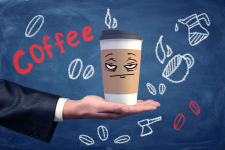 A businessmans hand holding a coffee cup with a drawn unhappy grumpy face on it.