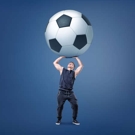 A muscular man tries to hold a giant and heavy black and white football ball over himself.の写真素材