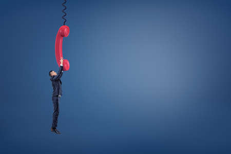 A small businessman holds a giant red retro phone handle and hangs from it trying not to fall.の写真素材