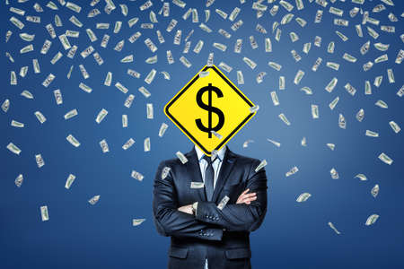 A businessman stands under a rainfall of dollar banknotes with a dollar road sign instead of his head.の写真素材