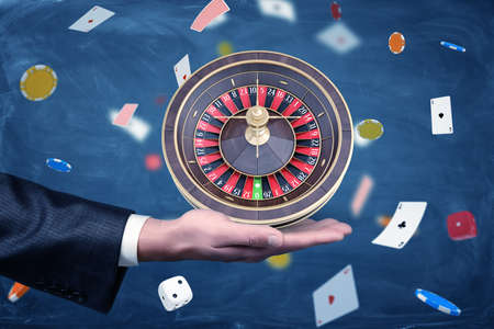 A businessman holds a casino roulette on his palm on a background of chips and cards.