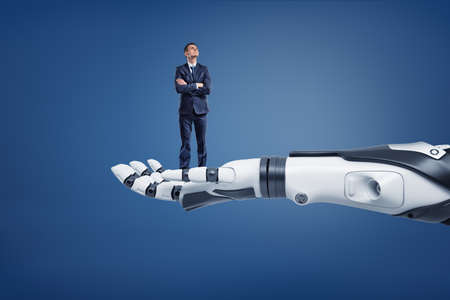 A small thinking businessman looks up while standing on a huge robotic arm.の写真素材