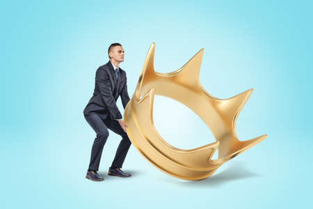 A young businessman in a smart suit trying to lift a huge golden crown from the floor.の写真素材