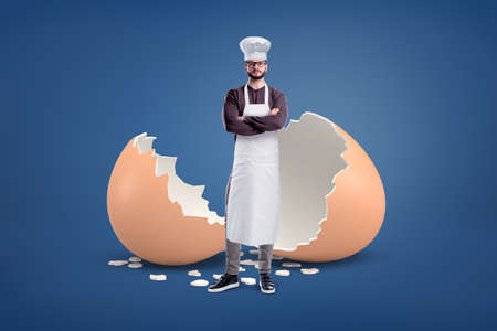 A young bearded cook in a white apron and a hat stands near a large broken egg shell.の写真素材
