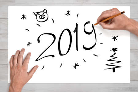 Male hands holding pencil above white sheet of paper with 2019 sign, pig face, christmas tree and snowflakes drawn onの写真素材