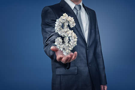 A businessman with unseen face shows an open palm with a dollar sign made of many paper banknotes.