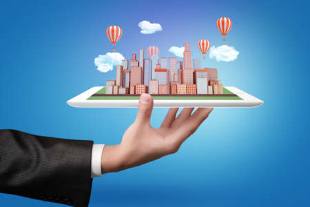 Businessman hand holding a tablet with city skyscrapers model, clouds and air balloons on blue...