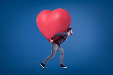 A side view of a young handsome man in a casual outfit carrying a huge red heart on his back.の写真素材