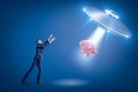 A businessman raising hands toward a UFO which is pulling a piggy bank toward its open hatch by some...