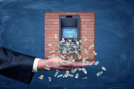 A businessmans palm holding a piece of a brick wall section with an ATM machine giving out dollar...