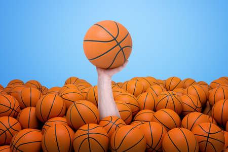 Hand appearing out of heap of orange basketball balls holding one on blue backgroundの写真素材
