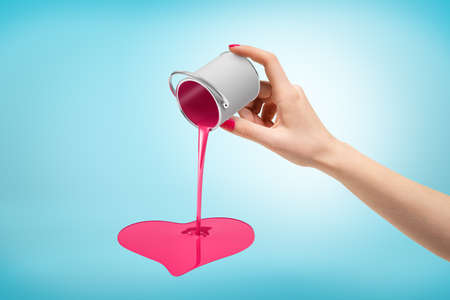 Female hand turning small silver paint bucket upside down with pink heart shaped pouring on blue...