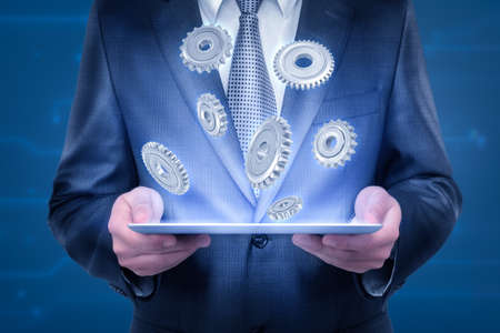 Crop close-up of businessmans chest and hands holding thin tablet lighting up some cogwheels flying above it.の写真素材