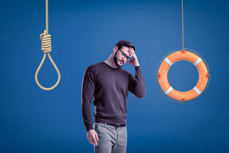 Man in casual outfit standing half-turn with hand on forehead as if choosing between lifebuoy hanging on rope and hangmans noose.の写真素材