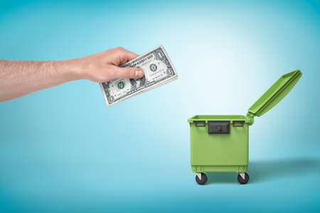 Male hand throwing bundle of money dollars into green trash bin on blue backgroundの写真素材