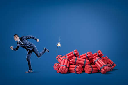 Businessman running away from pack of red tnt dynamite sticks with lighted fuse on blue background. Management and marketing. Financial risks. Business and commerce.の写真素材