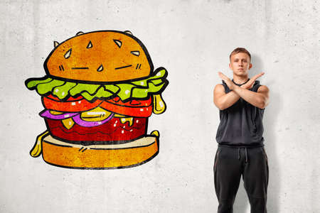 Young fit man in sleeveless hoody standing with arms crossed across chest at white wall with drawing of huge hamburger on it.の写真素材