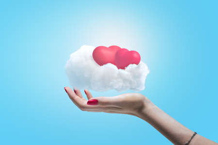 Female hand holding white cloud with two pink hearts on blue sky background