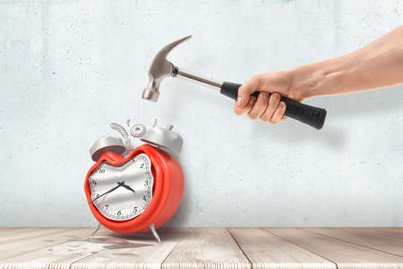 A mans hand holding a hammer and crashing a retro alarm clock standing on the wooden floor.の写真素材