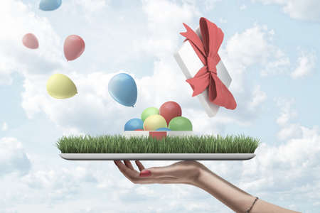 Side view of womans hand holding tray owith patch of green lawn with open gift box on top from which...