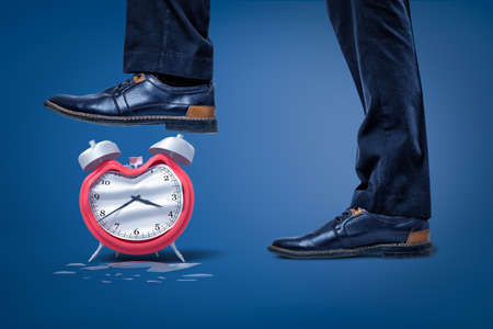Side close-up of mans feet, one foot raised to crush broken bent alarm clock.