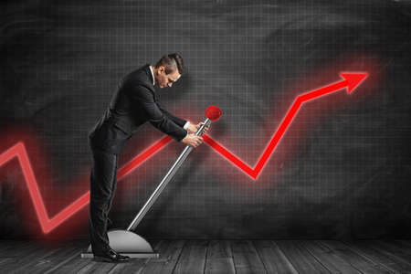 Side view of businessman looking down and pulling big lever near black graph ruled wall with red graph arrow on it.の写真素材