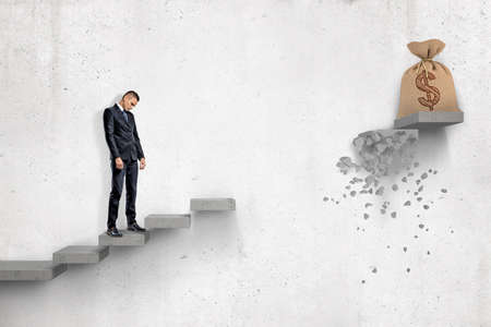 Sad businessman looking down on gray concrete staircase with big money bag at the top of it but with...