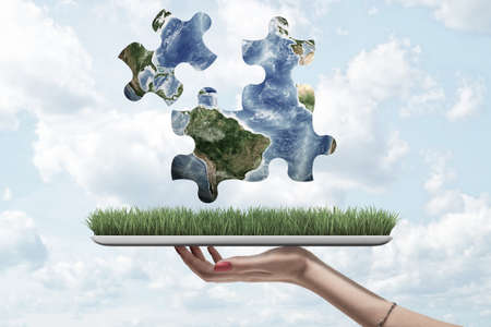 Female hand holding a grass model and earth globe puzzle pieces above on blue sky background