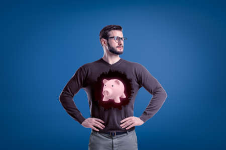 Man with glasses and a pink piggy bank on a grey jumper on blue backgroundの写真素材
