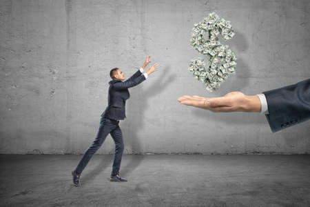 Side view of miniatured businessman holding out hands to grab dollar symbol formed from many banknotes suspended in air above big mans hand.の写真素材